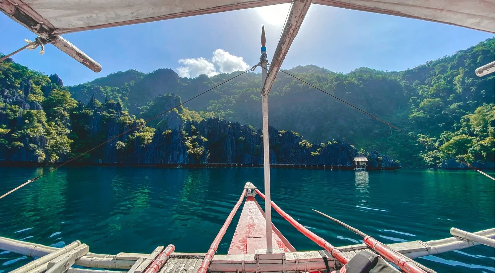 View from the bow of an outrigger boat heading across calm green water toward forested limestone cliffs