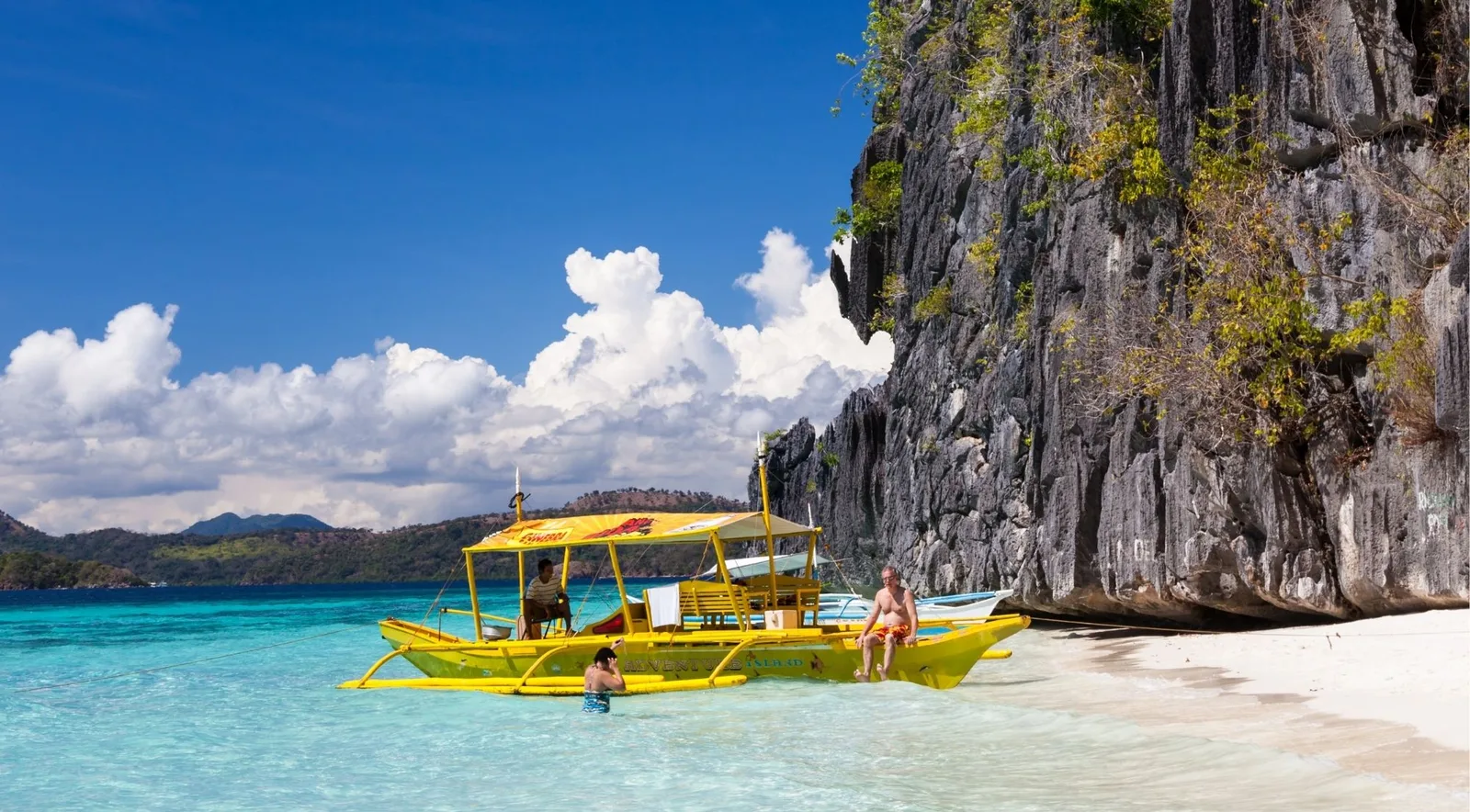 A yellow outrigger boat pulled up on a white-sand beach beside a limestone cliff, with swimmers in the shallows