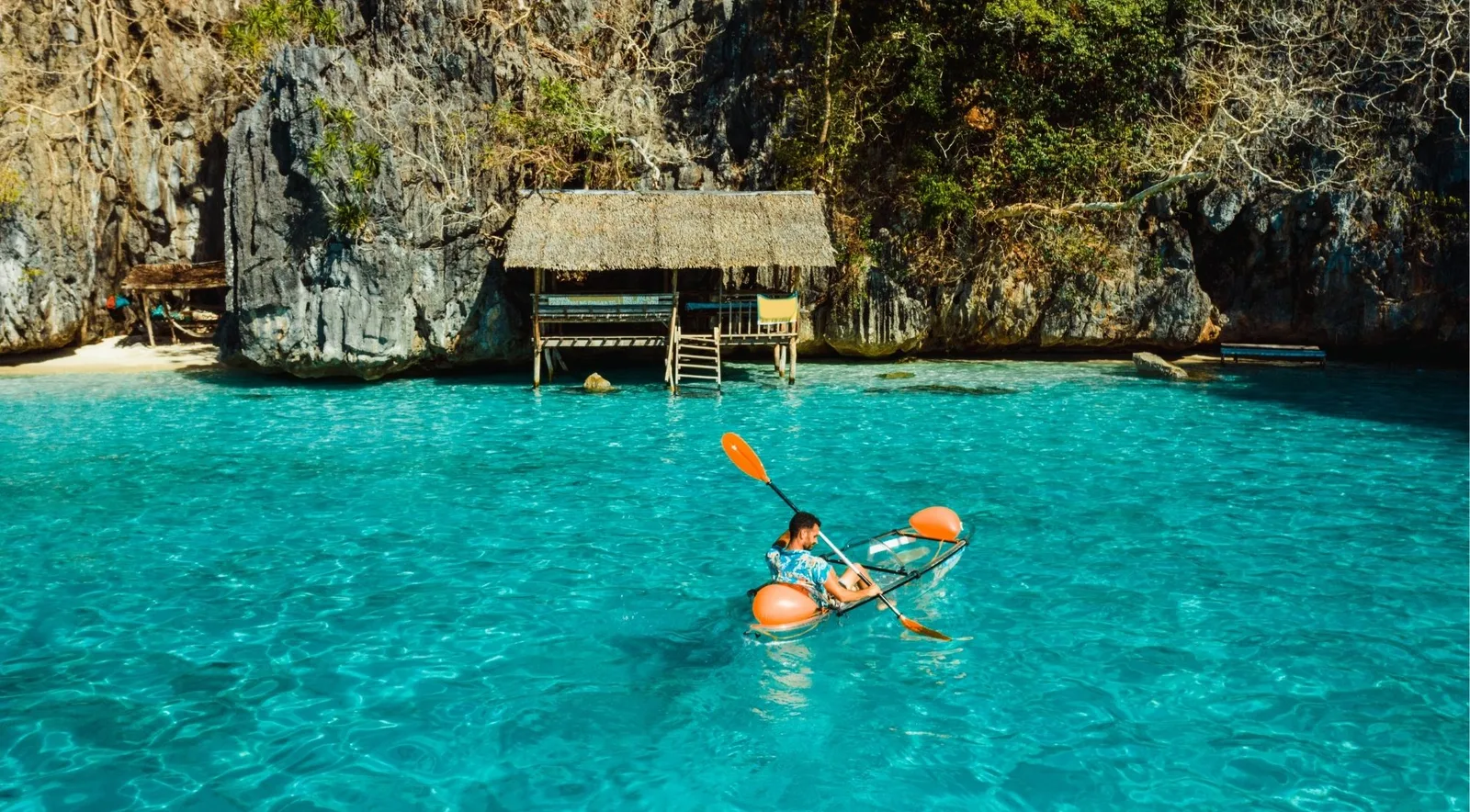 A kayaker paddling across clear turquoise water toward a thatched hut on stilts at the foot of a limestone wall