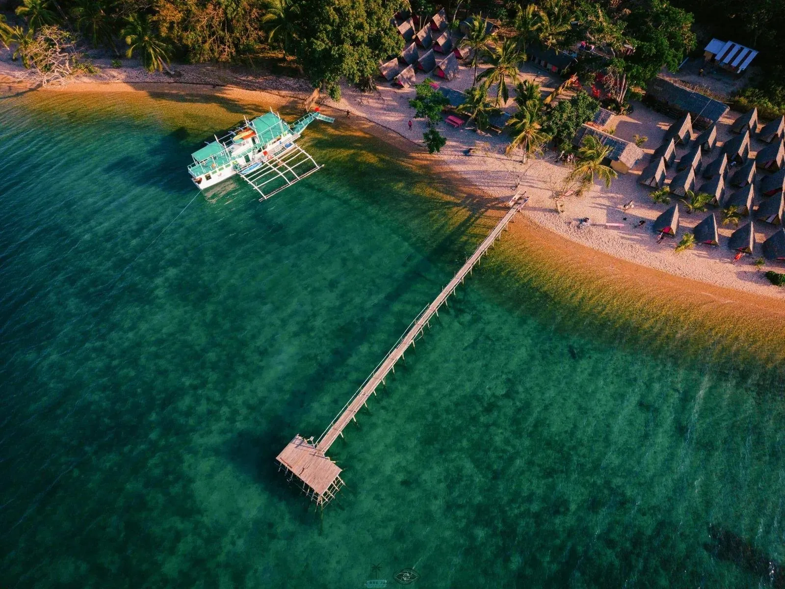 Aerial view of a beach camp with a long wooden jetty and an anchored outrigger boat