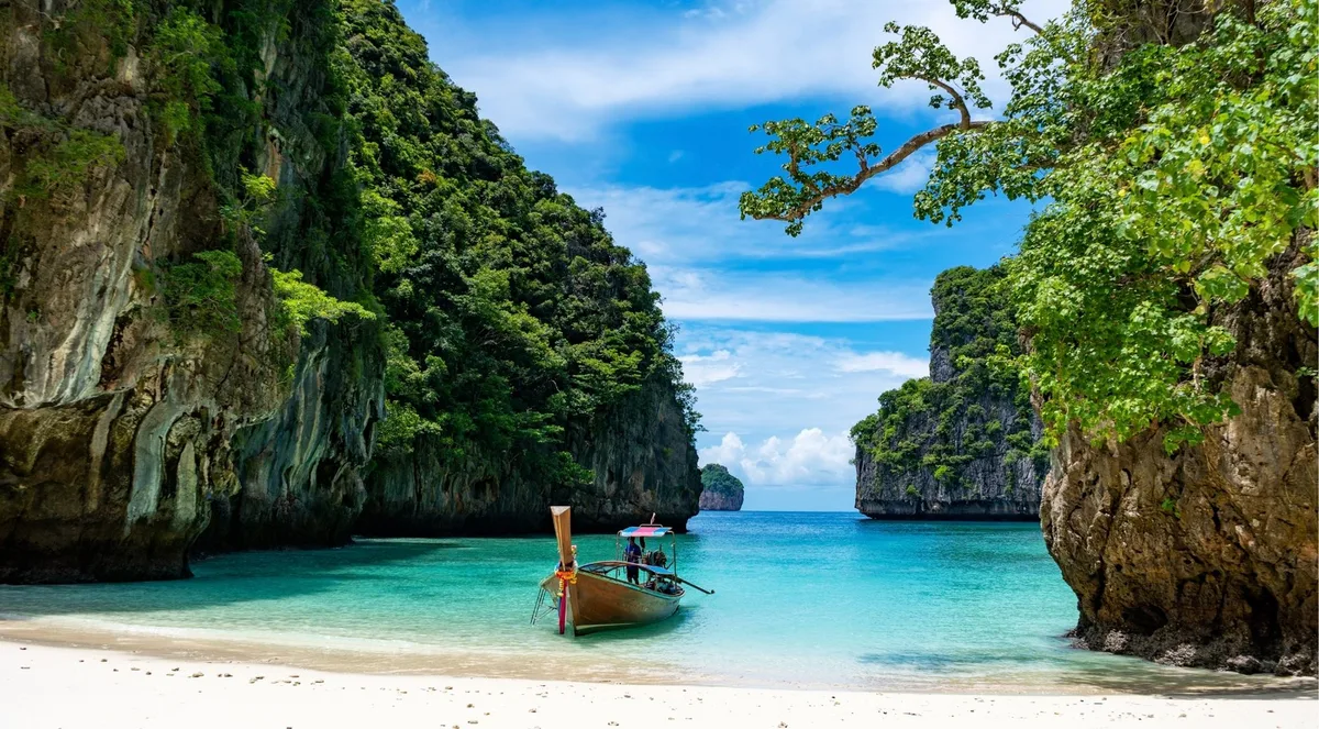 A wooden boat anchored in turquoise shallows off a white-sand beach, between towering limestone cliffs