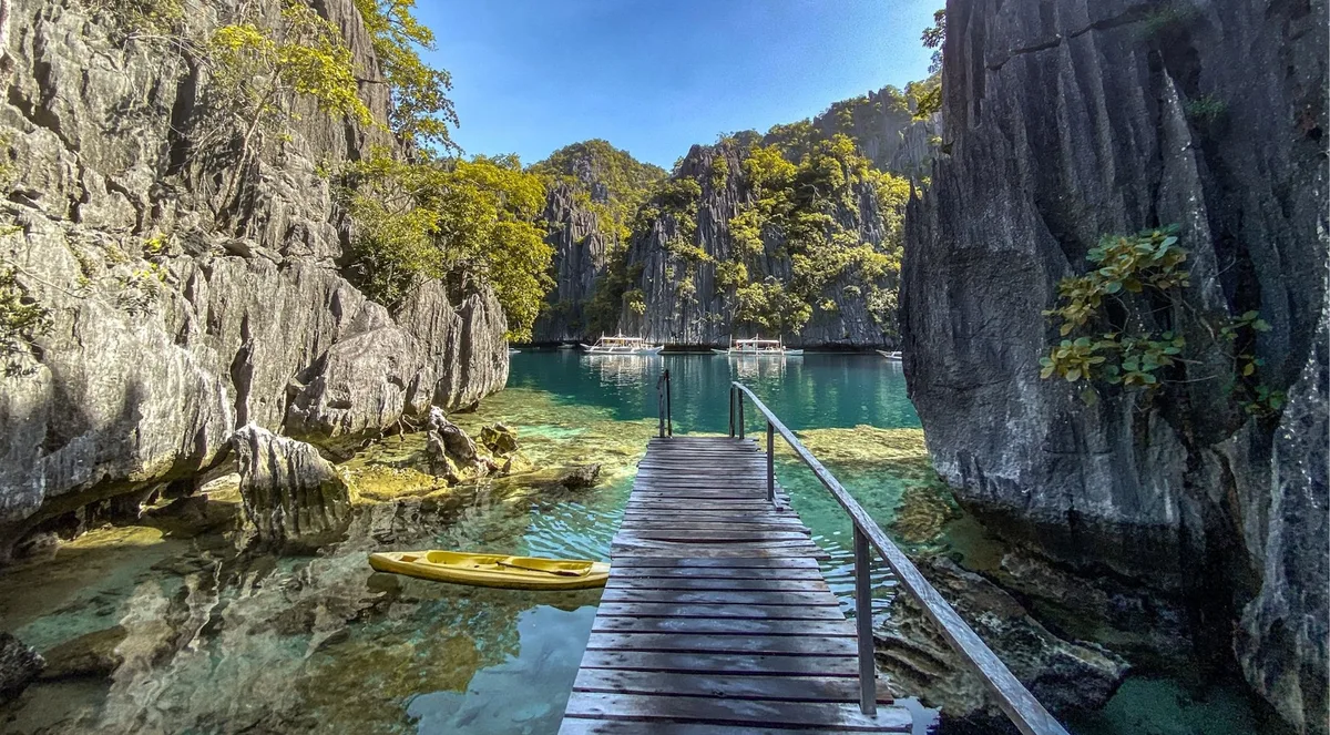 A wooden boardwalk leading out into a clear green lagoon between steep limestone walls, with a kayak moored at the end