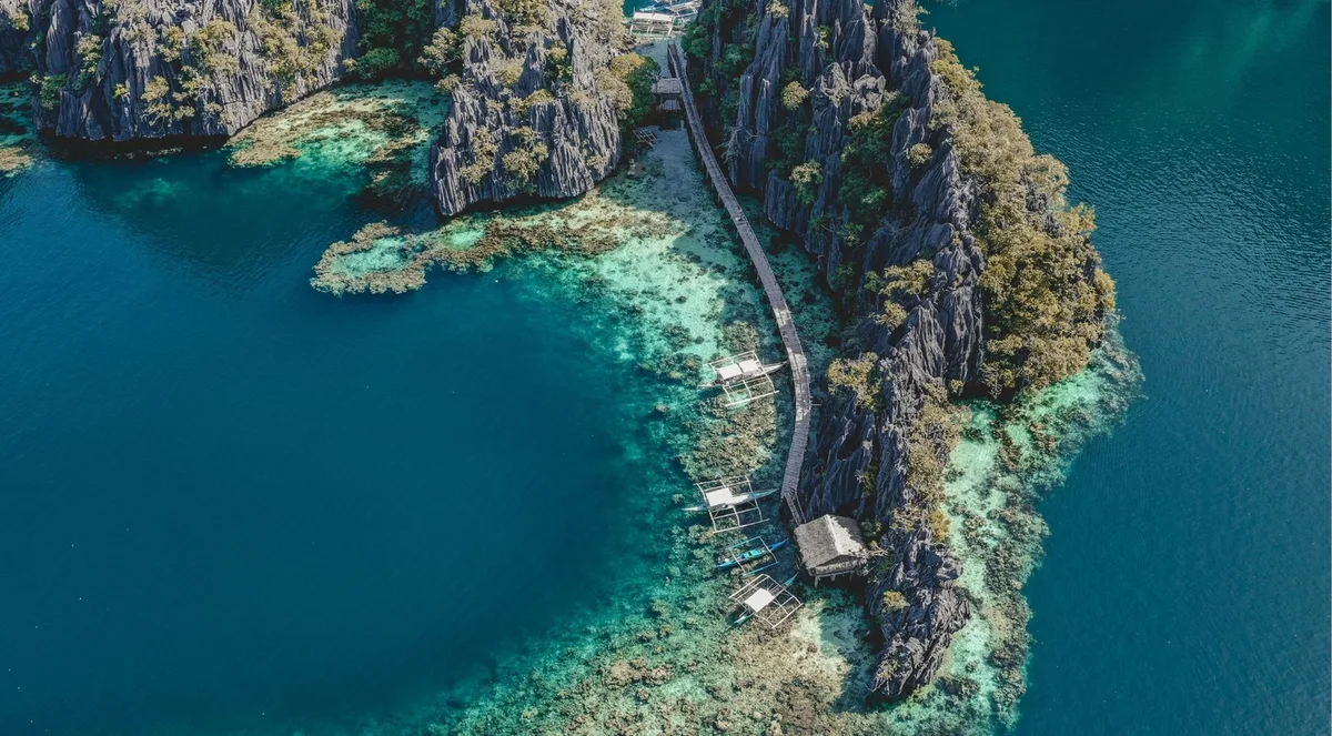 Aerial view of a wooden boardwalk running along the edge of a limestone island, with stilt huts standing over turquoise water