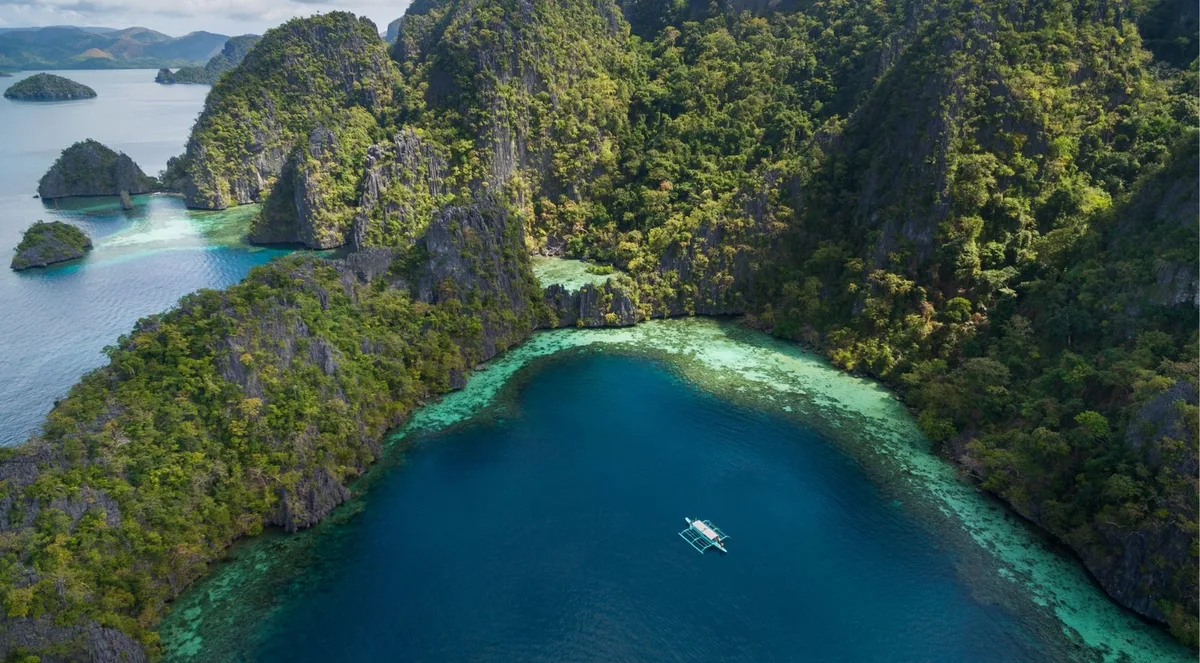 Aerial view of forested limestone ridges around a deep-blue lagoon with a small boat on the water, Coron Island