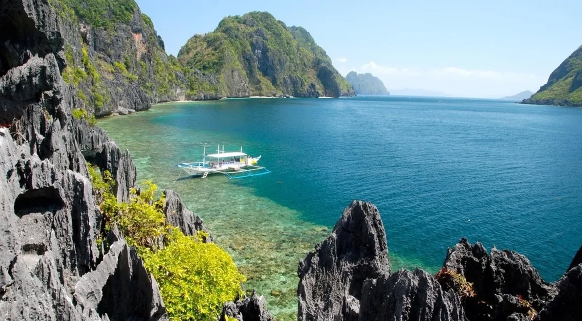 View over dark limestone rocks to a calm bay with outrigger boats moored offshore, El Nido