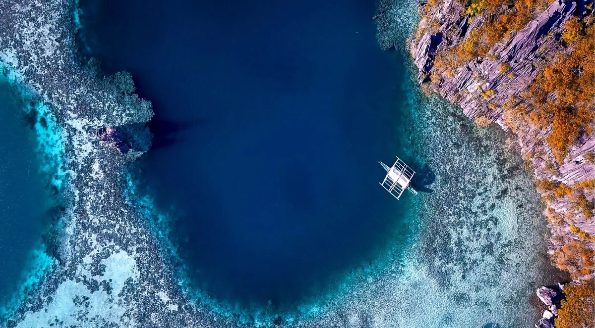 Aerial view of a deep-blue hole in pale reef shallows beside limestone cliffs, with a single outrigger boat on the water