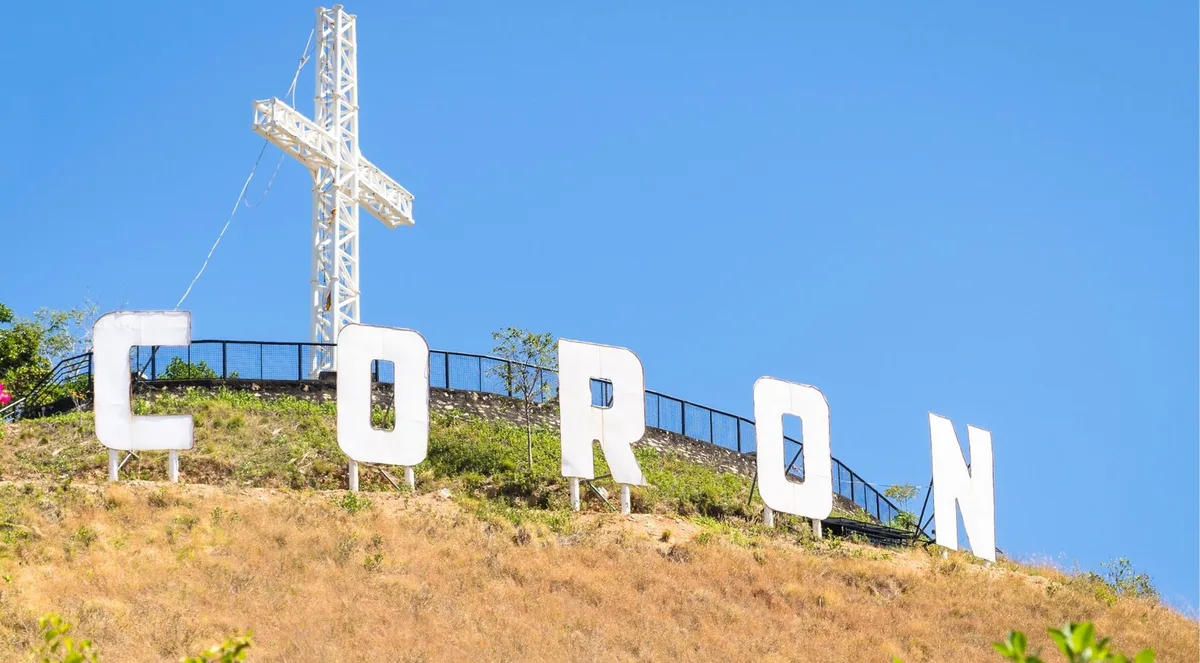 Hilltop with giant white CORON letters and a cross above the roofs of Coron town and the bay