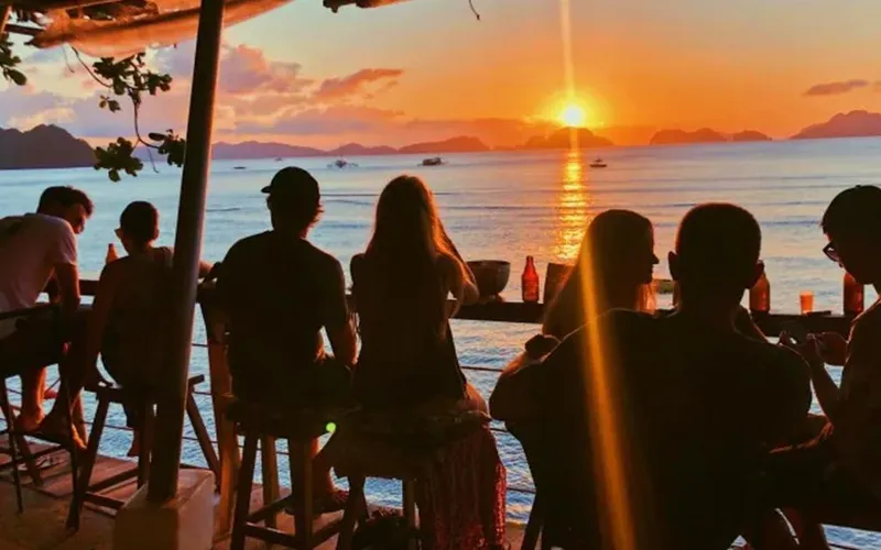 Guests at the bar of Outpost Beach Hostel watching the sun set over the bay